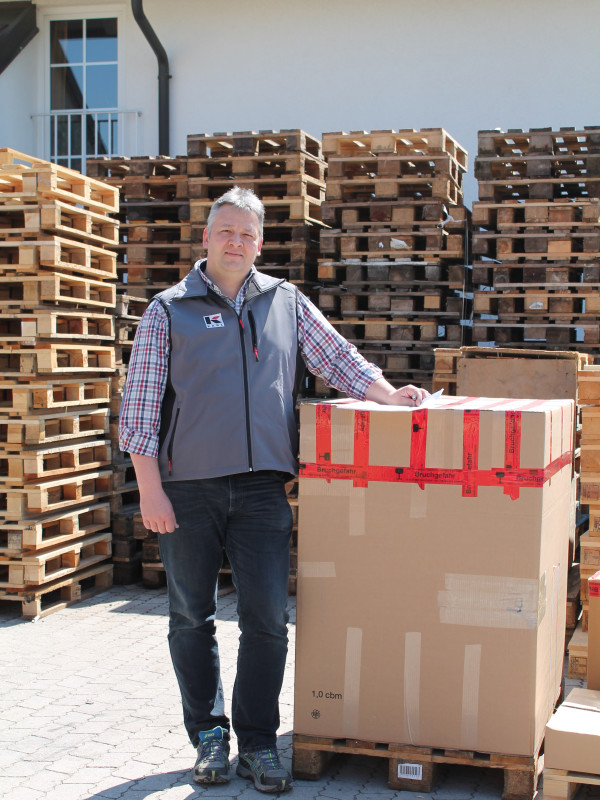 Mann steht neben einem großen Karton auf einer Palette, umgeben von gestapelten Holzpaletten in einem Lagerbereich im Freien.