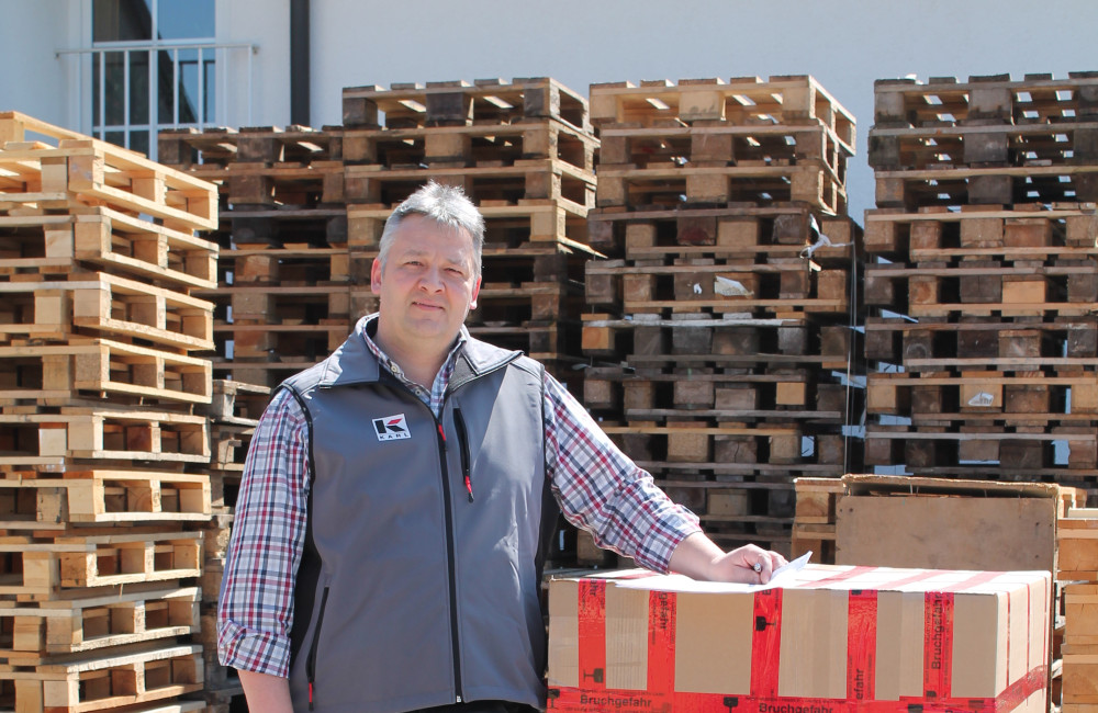 Mann steht neben einem großen Karton auf einer Palette, umgeben von gestapelten Holzpaletten in einem Lagerbereich im Freien.