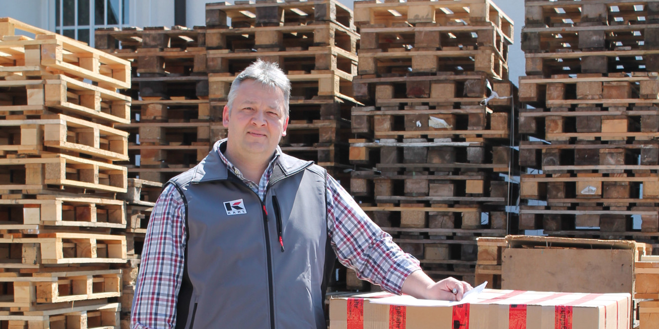 Mann steht neben einem großen Karton auf einer Palette, umgeben von gestapelten Holzpaletten in einem Lagerbereich im Freien.